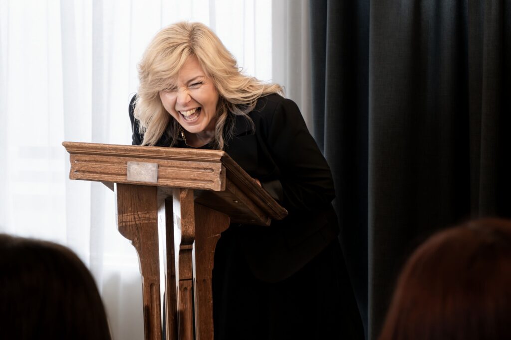 Blonde woman stands at a lectern laughing | It's okay to laugh at a funeral